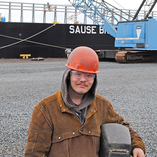young man standing in front of a cargo ship wearing welding gear