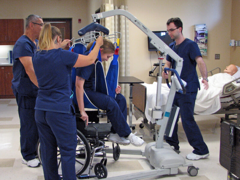 curry nurses_3420 Image of 4 Curry Campus nursing students in a lab practicing a lift assist with a medical apparatus