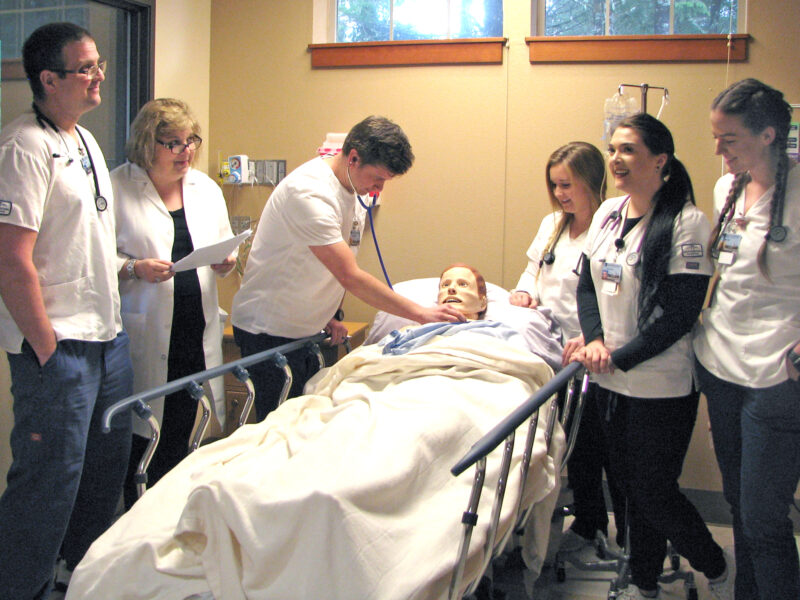 curry nurses_3896 Image of 5 Curry Campus nursing students and an instructor standing around a patient simulator (in a hospital bed) in the Nursing Lab