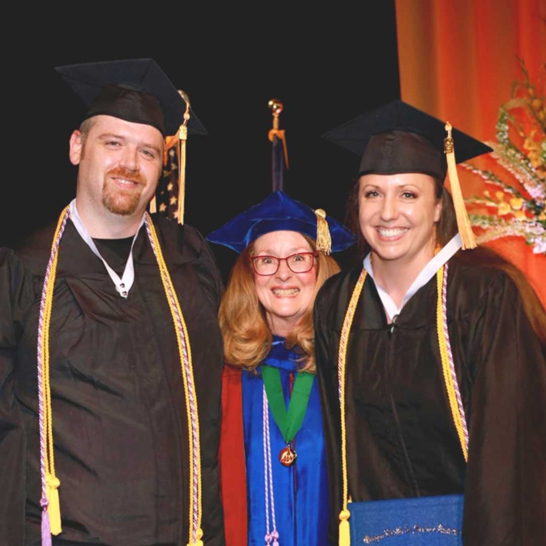 Three people standing on a stage graduating wearing regalia including graduation hats; Male student (Harley) on the left, distinguished woman in middle, and female student (Heather) on the right holding a diploma