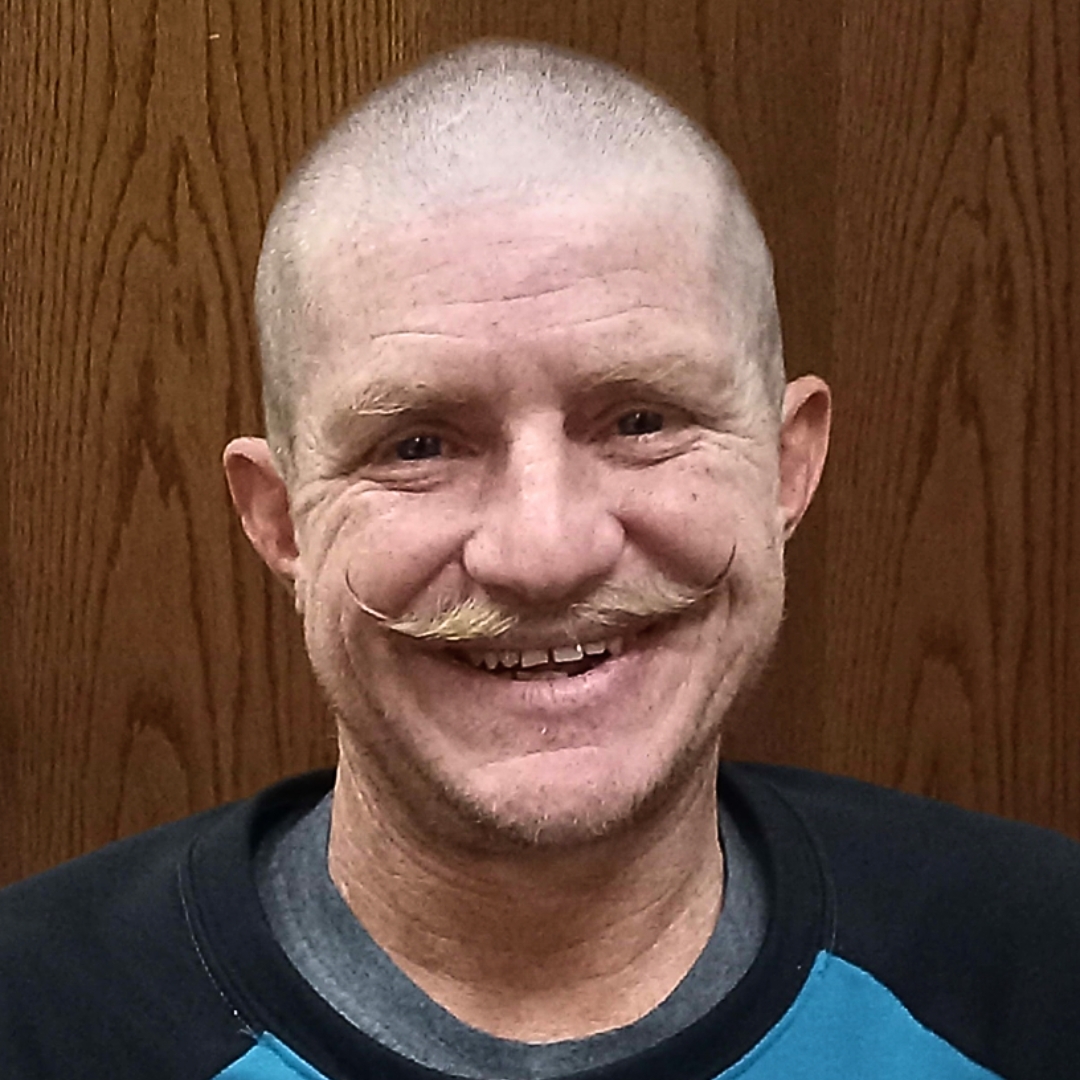 Profile image of male student with mustache smiling and standing in front of wooden background