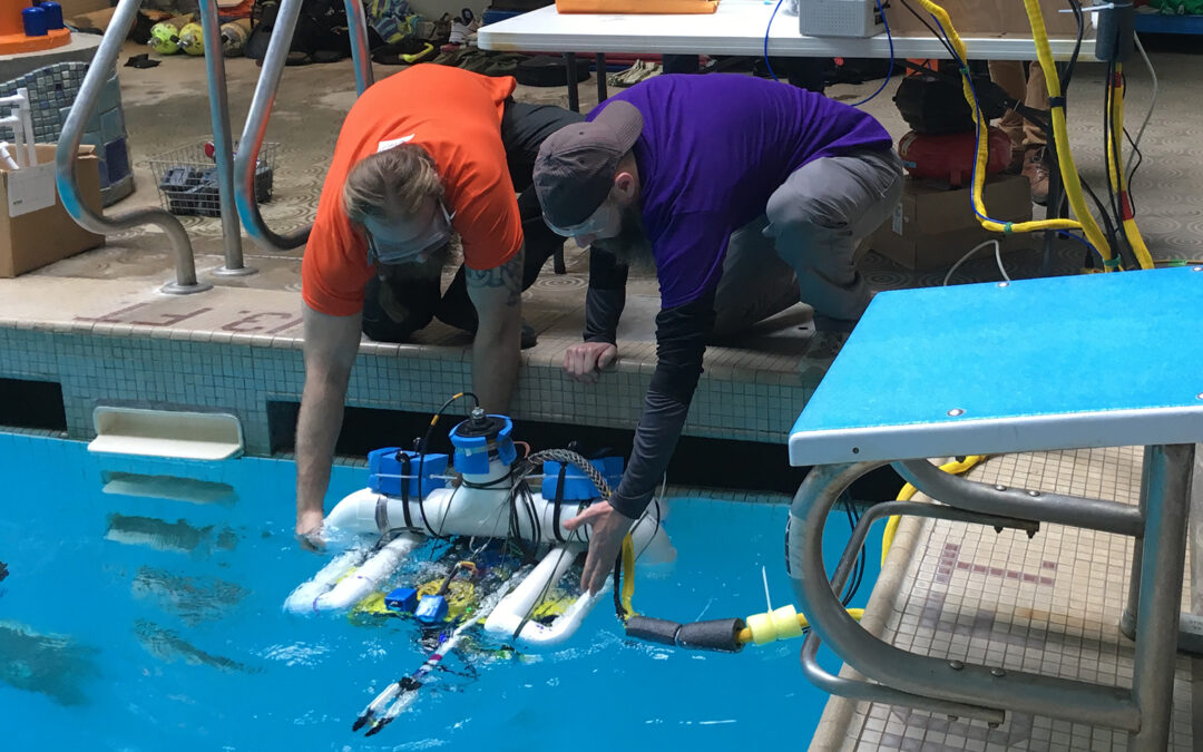 Image is of two people lowering a remote operated vehicle into a swimming pool