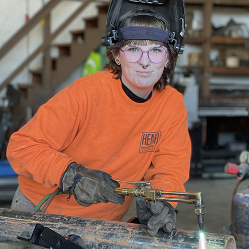 Image is a woman wearing a welding helmet opened so you can see her smiling, she's holding a flaming torch on a pipe