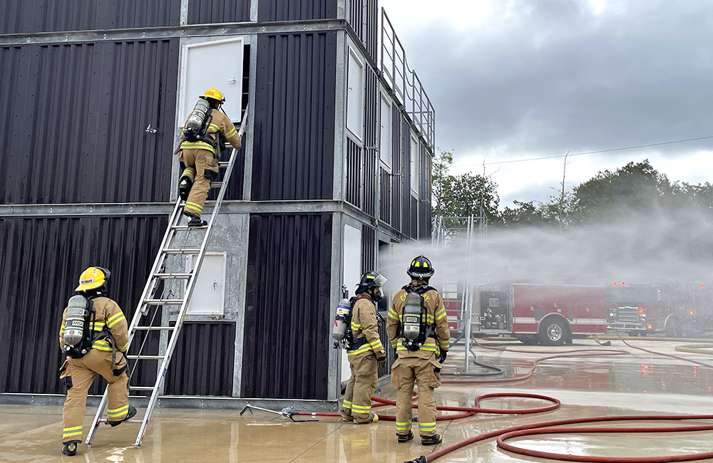 Image is of four firefighters around a fire training tower, one is on a ladder, there is smoke and water all around. There is a fire truck in the background.