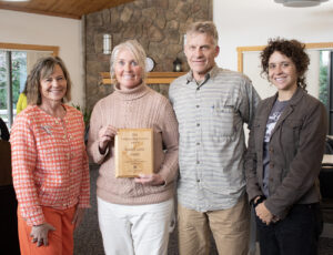 Image is a group of four people standing side by side smiling. One is holding a plaque.