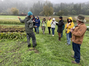 Image is a group of people in a circle listening to a woman speak, background is a farm scene