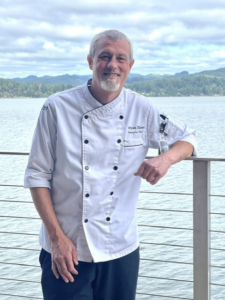 Portrait of Chef Bryan Elam standing outdoors leaning on a railing with a bay view in the background