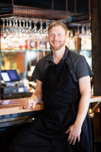 Portrait of Chef Rob Chester standing in a restaurant in front of a bar with wine glasses hanging in the background