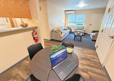 image of the dining area of an apartment showing dining table, four chairs, laptop computer, looking across the room into the living room area