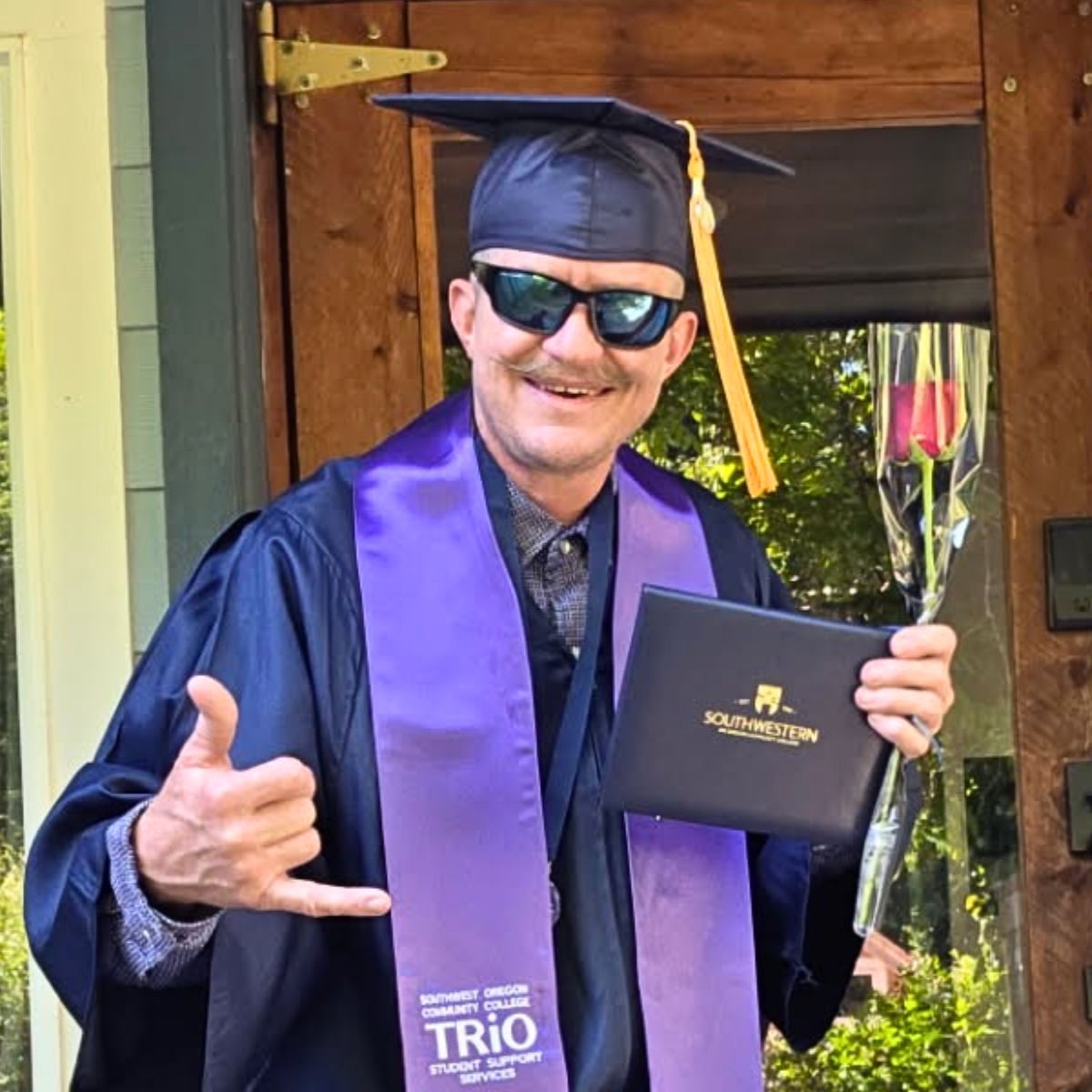 Profile image of STEP graduate, Jonathan, standing in front of a door in graduation regalia holding a rose and a diploma