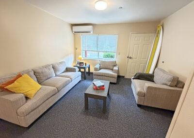 image is of the apartment living room showing the couch, chairs and two coffee tables, door in the corner, one large window and a surfboard in the corner